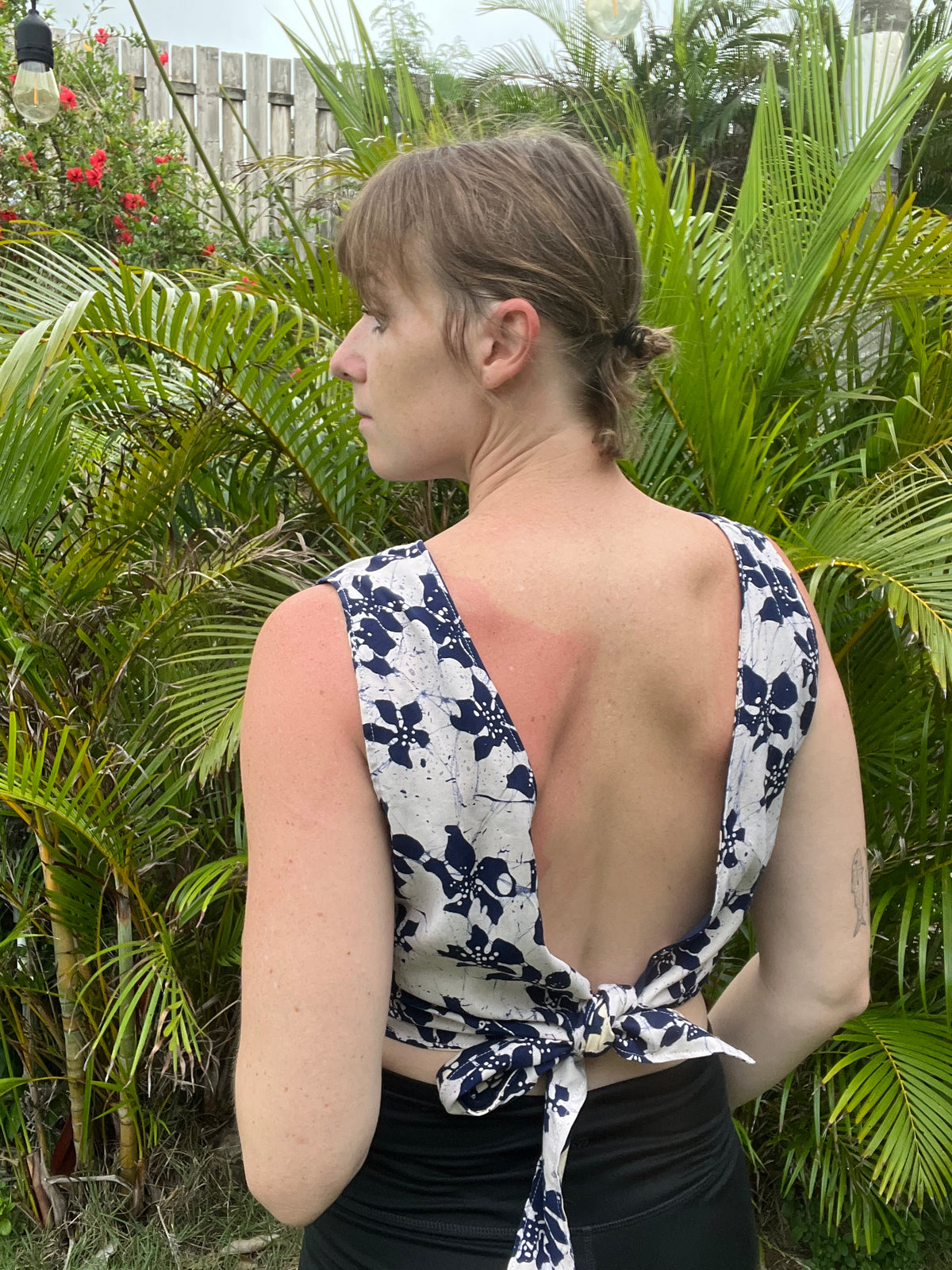 A woman shown from the back wearing a white sleeveless top with a navy floral print and an open back tied at the waist, standing outdoors in front of lush green tropical plants.