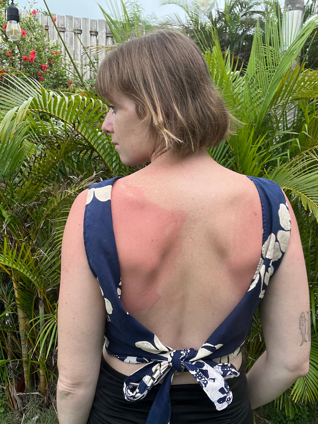 A woman shown from behind wearing a navy blue and white floral print open-back top with a tie detail at the waist, standing outdoors in front of lush green tropical plants.