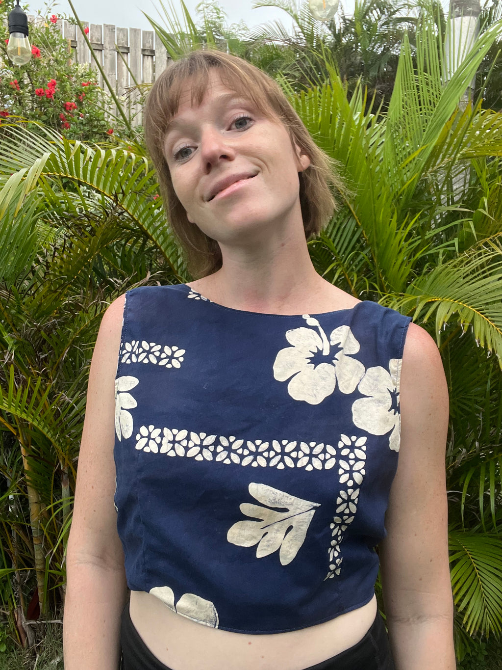 A woman wearing a navy blue sleeveless crop top with a white hibiscus floral and geometric pattern, standing outdoors in front of lush green tropical foliage.
