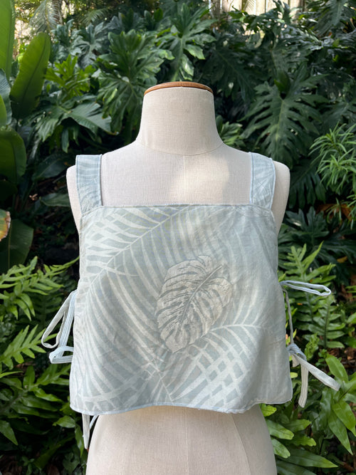 A sleeveless light blue top with a tonal palm leaf print and side tie details, displayed on a cream-colored dress form mannequin against a background of lush green tropical foliage.