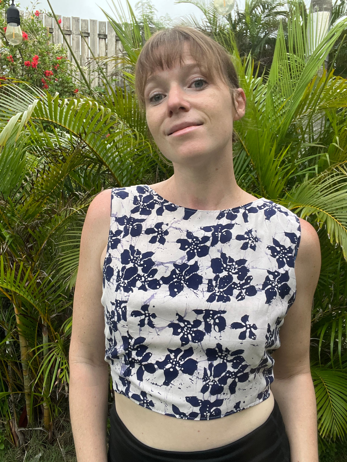 A woman wearing a sleeveless white crop top with a navy blue floral pattern, standing outdoors in front of lush green tropical foliage.