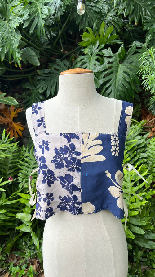 A sleeveless patchwork floral top displayed on a cream fabric mannequin torso, set against a lush background of green tropical foliage.
