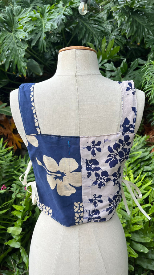 A patchwork-style fabric top displayed on a cream fabric mannequin torso, photographed outdoors in front of dense green foliage. The garment has navy and off-white floral panels with white side ties.